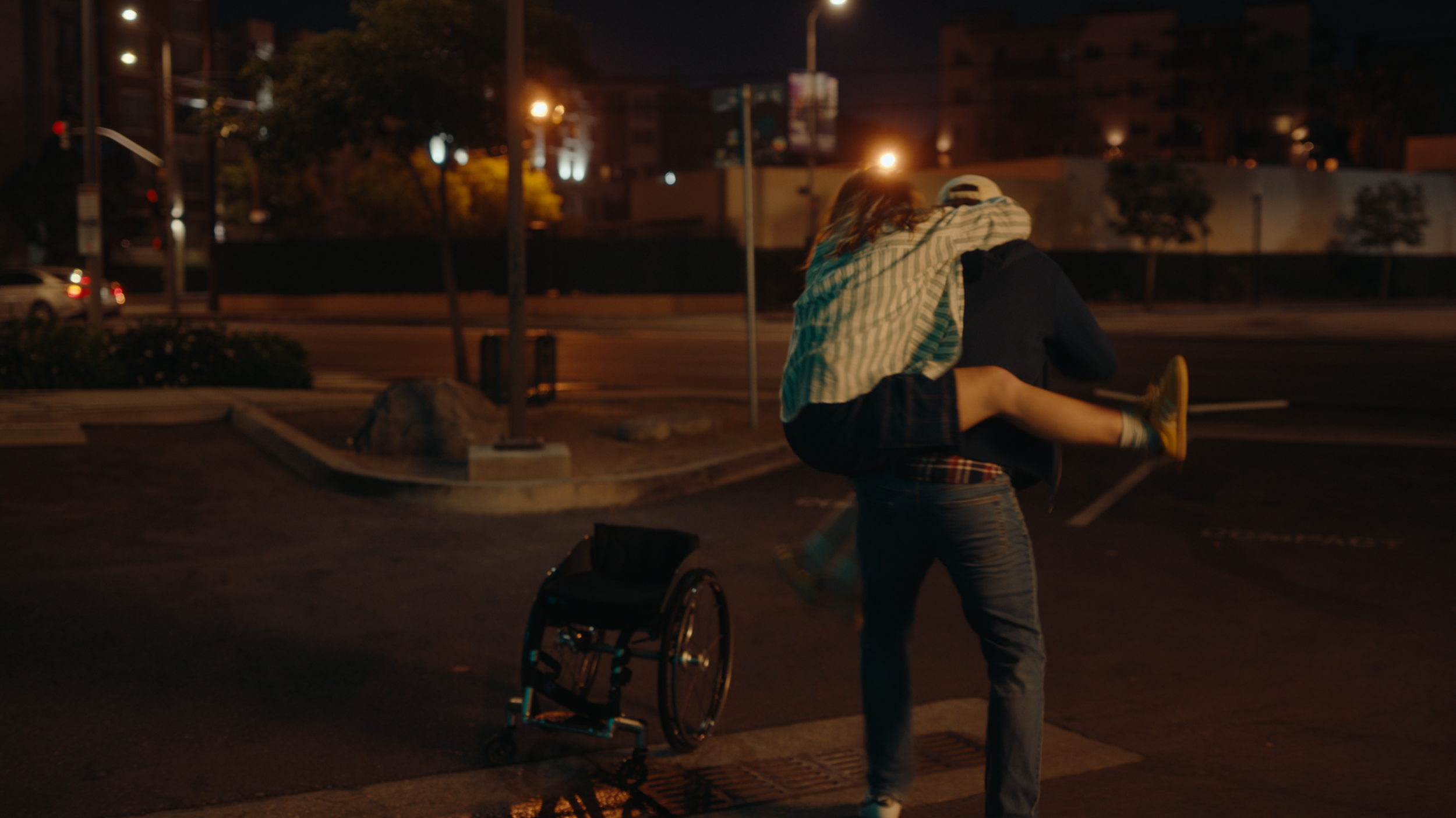 A person wearing a hat lifts a woman in a striped shirt in a parking lot at night. The parking lot has a rollator walker nearby, with city lights and trees in the background.