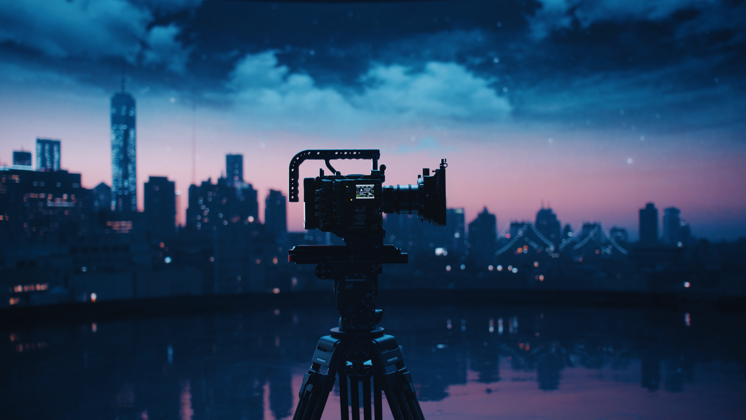 Camera on tripod facing city skyline at dusk with clouds and lit buildings.