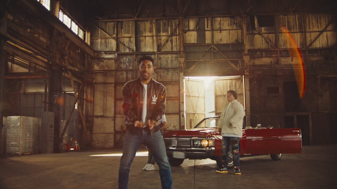 Three men standing inside a rustic, industrial warehouse with a red vintage car in the background. The man in the foreground is wearing a black jacket with patches, a white shirt, and jeans. The man to the right wears a light-colored jacket and jeans, standing near the open car door. The third man is partially visible behind the man in the foreground.