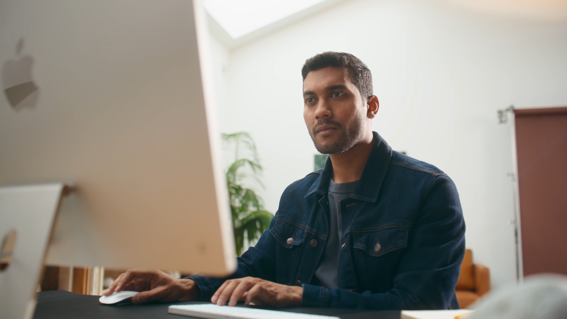 Man working on a computer with a focused expression in a bright office with a large window and green plants.