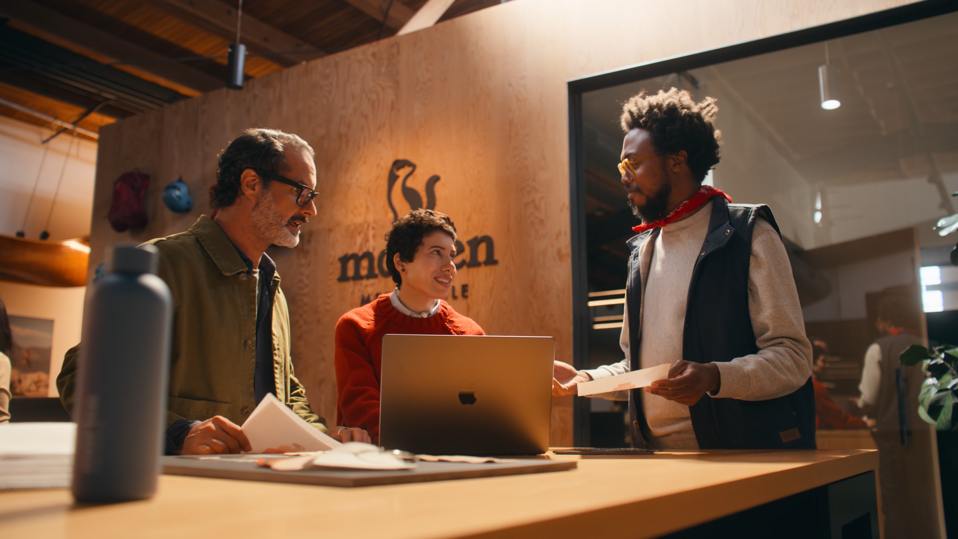 Three people at a modern reception desk, two men and a woman, engaging in conversation. One man is holding papers, the woman is smiling at a laptop, and the other man is handing papers to the woman. The setting appears to be a casual office or coworking space with wooden walls and a logo of a squirrel on the background wall.
