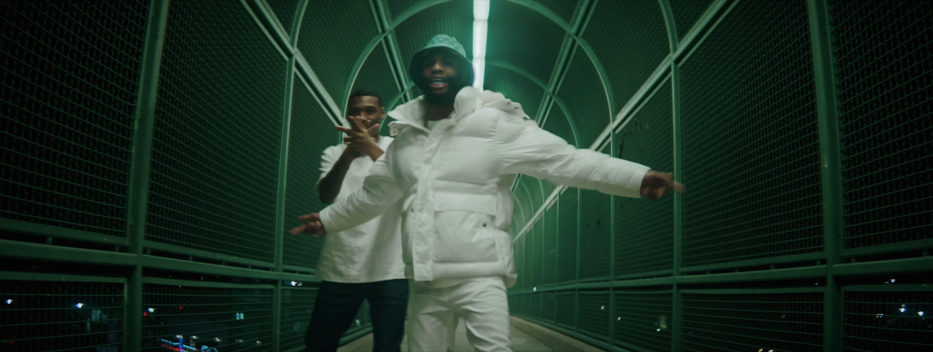 Two men in white clothing posing on a green enclosed bridge with metal railings at night.