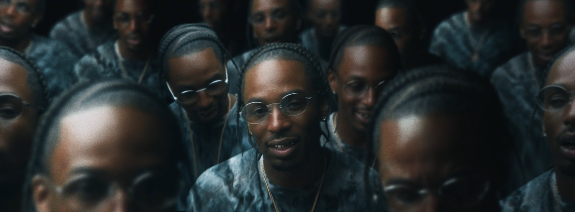 A group of people with braided hair and glasses, wearing matching outfits, closely gathered and looking down at the camera.