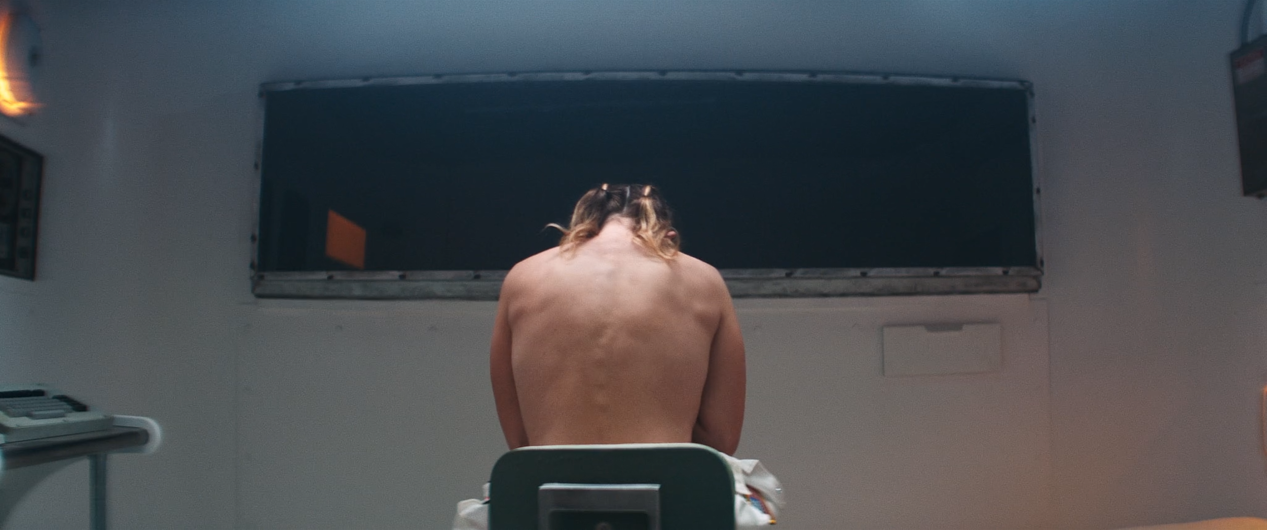 Person with bare back sitting in a room, facing a large black screen or board on the wall, with a small orange object visible inside.