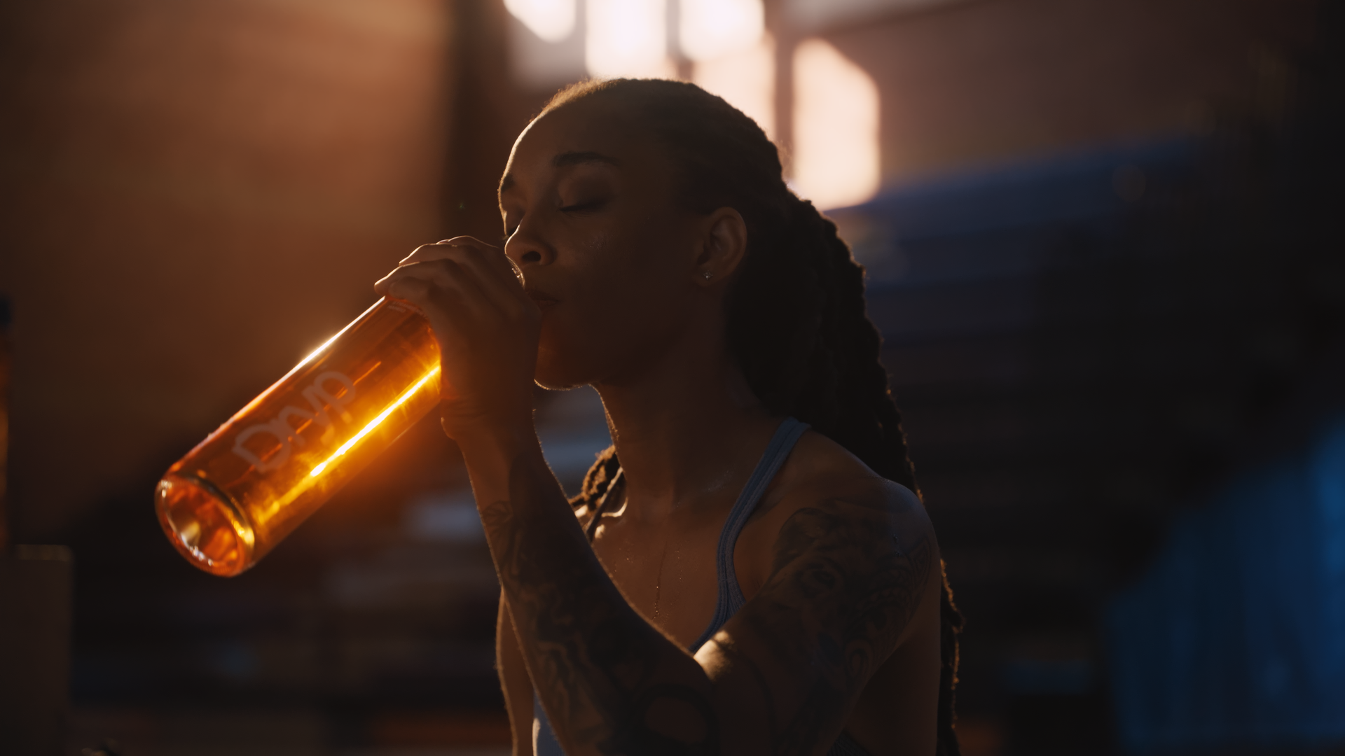 A woman with braided hair and tattoos on her arms is drinking from a transparent glass filled with an amber-colored beverage, in a dimly lit setting with sunlight streaming through a window in the background.