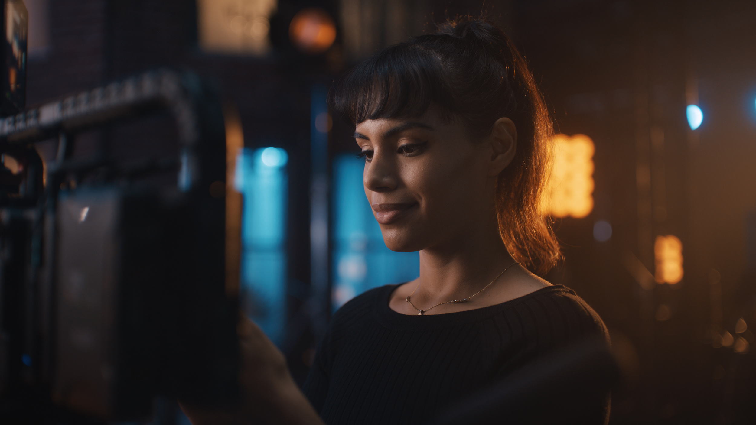 A young woman with short dark hair and a necklace looking at a screen in a dimly lit urban environment with blue and orange lights.