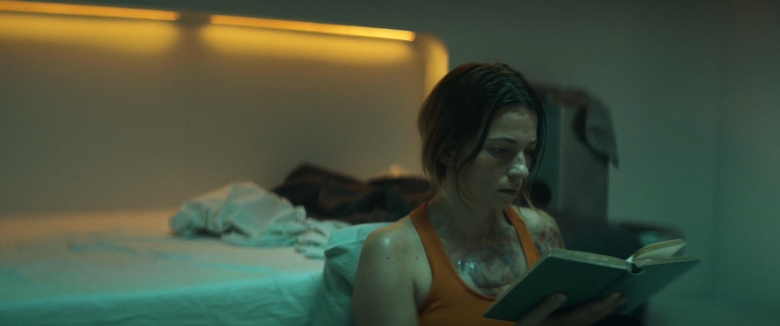 A woman with dark, wet hair and a dirty face sitting on a bed, reading a book in a dimly lit room.