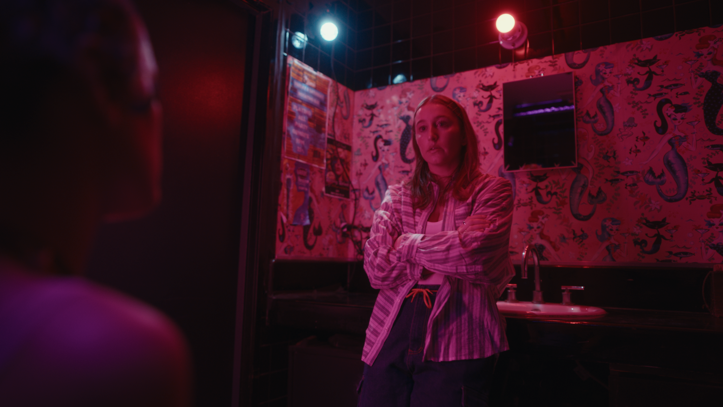 A woman with red hair and a striped shirt crossing her arms in front of a mirror in a pink-lit tattoo parlor with mermaid wallpaper.