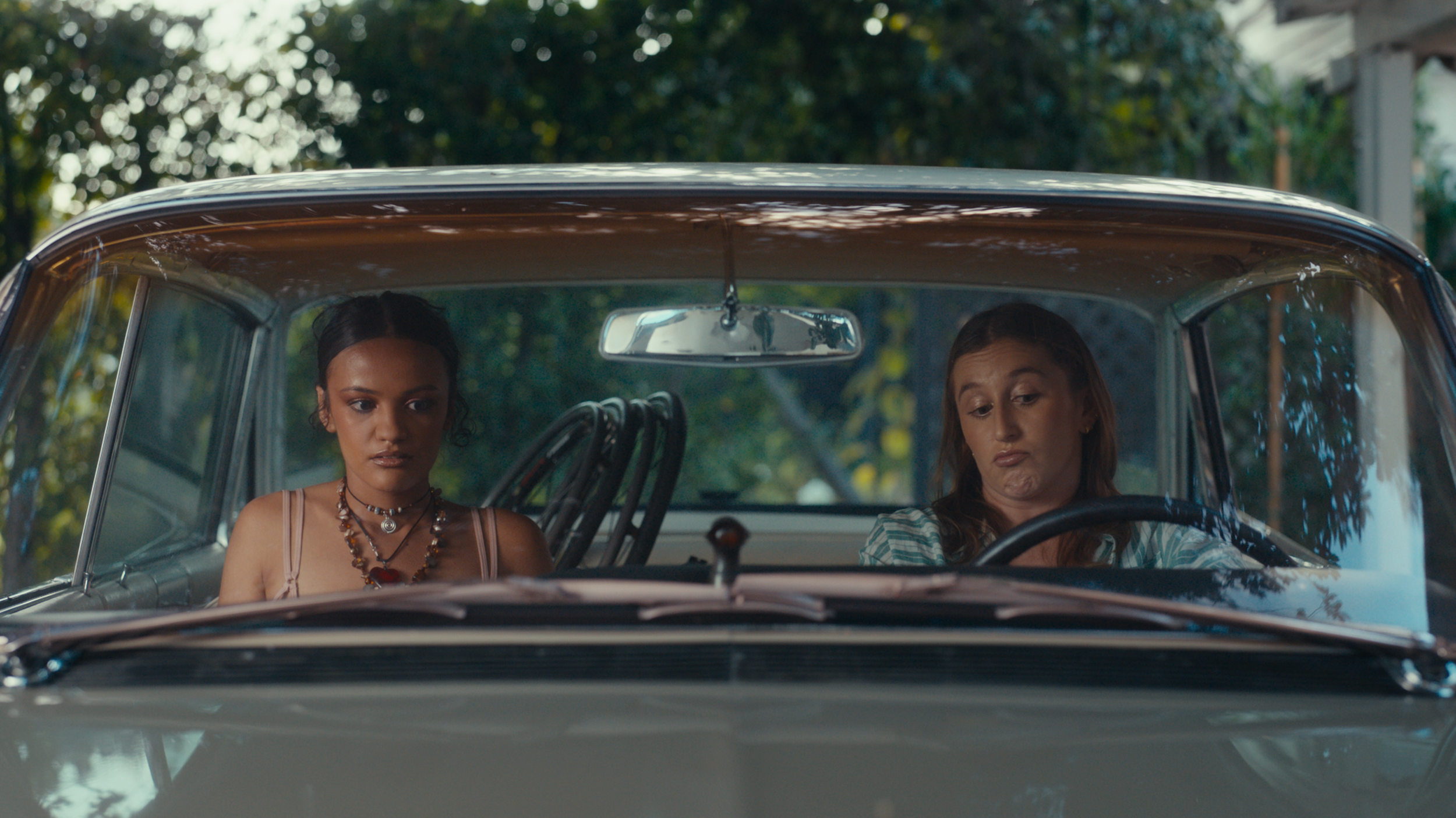 Two women sit in the front seats of a classic car, one driving with a serious expression and the other looking down with a neutral face. They are outdoors with trees in the background.