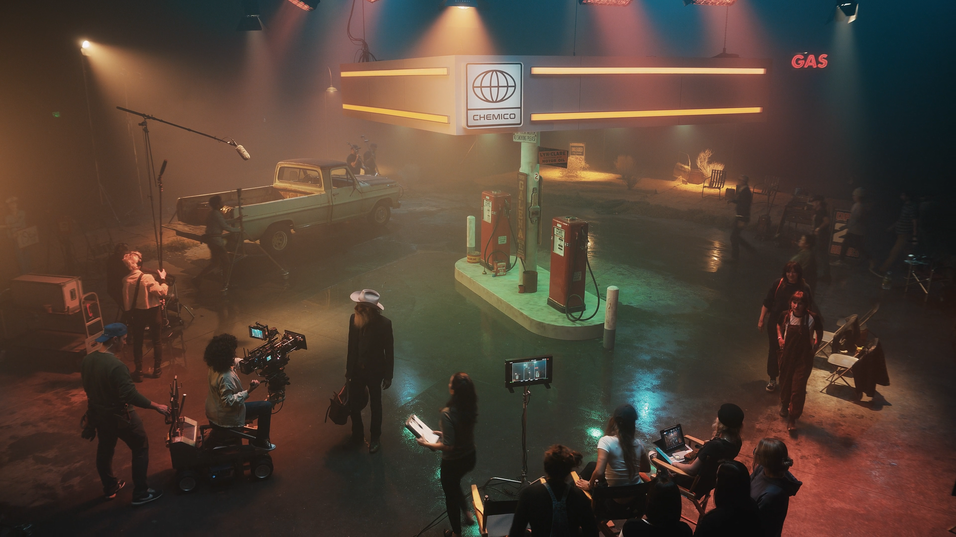 Indoor set with vintage gas station, old truck, and props for a filming or theatrical production, with crew members and equipment.
