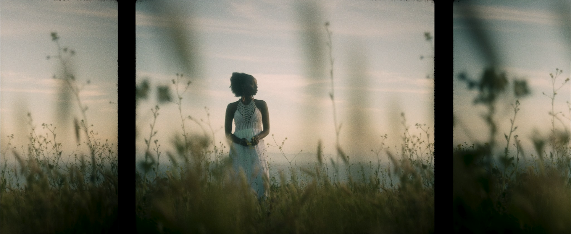 A woman in a white dress with lace details standing in a field with tall grasses and wildflowers, looking to the side, with a hazy sky in the background. The image appears to be viewed through a three-panel window or frame.