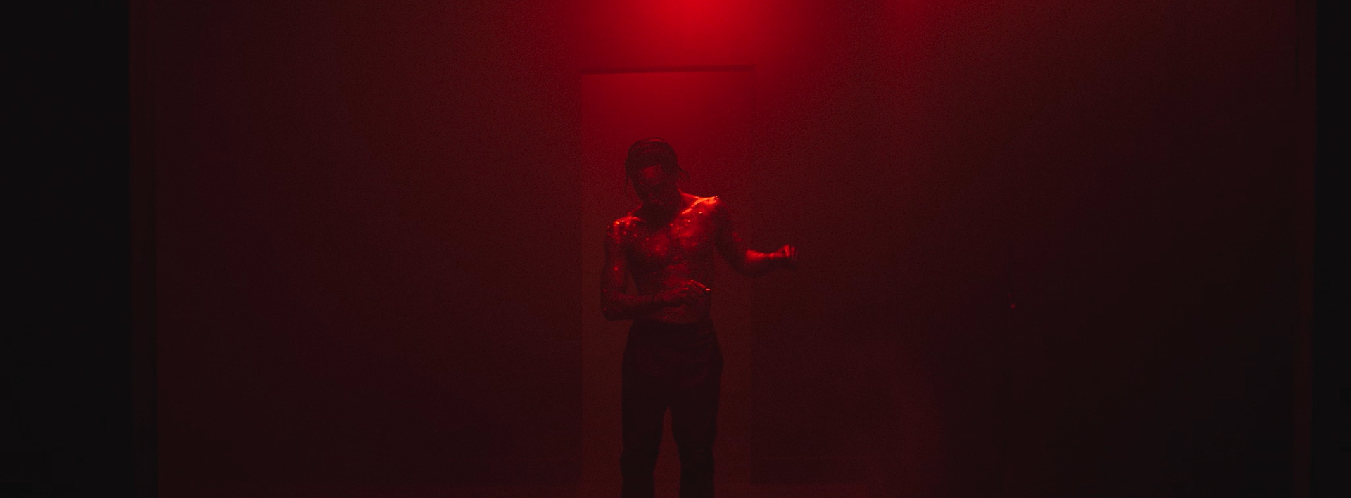 A person standing in a dark room illuminated by red light, with a textured or patterned shirt, wearing glasses, and appearing to be in a dance or expressive pose.