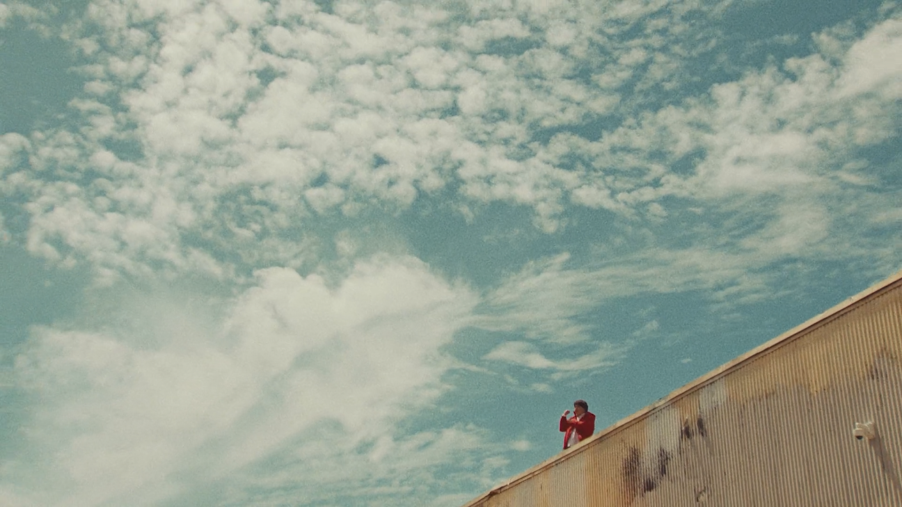 A person in a red coat taking a photo or video with a smartphone from the top of a large, weathered building against a partly cloudy sky.