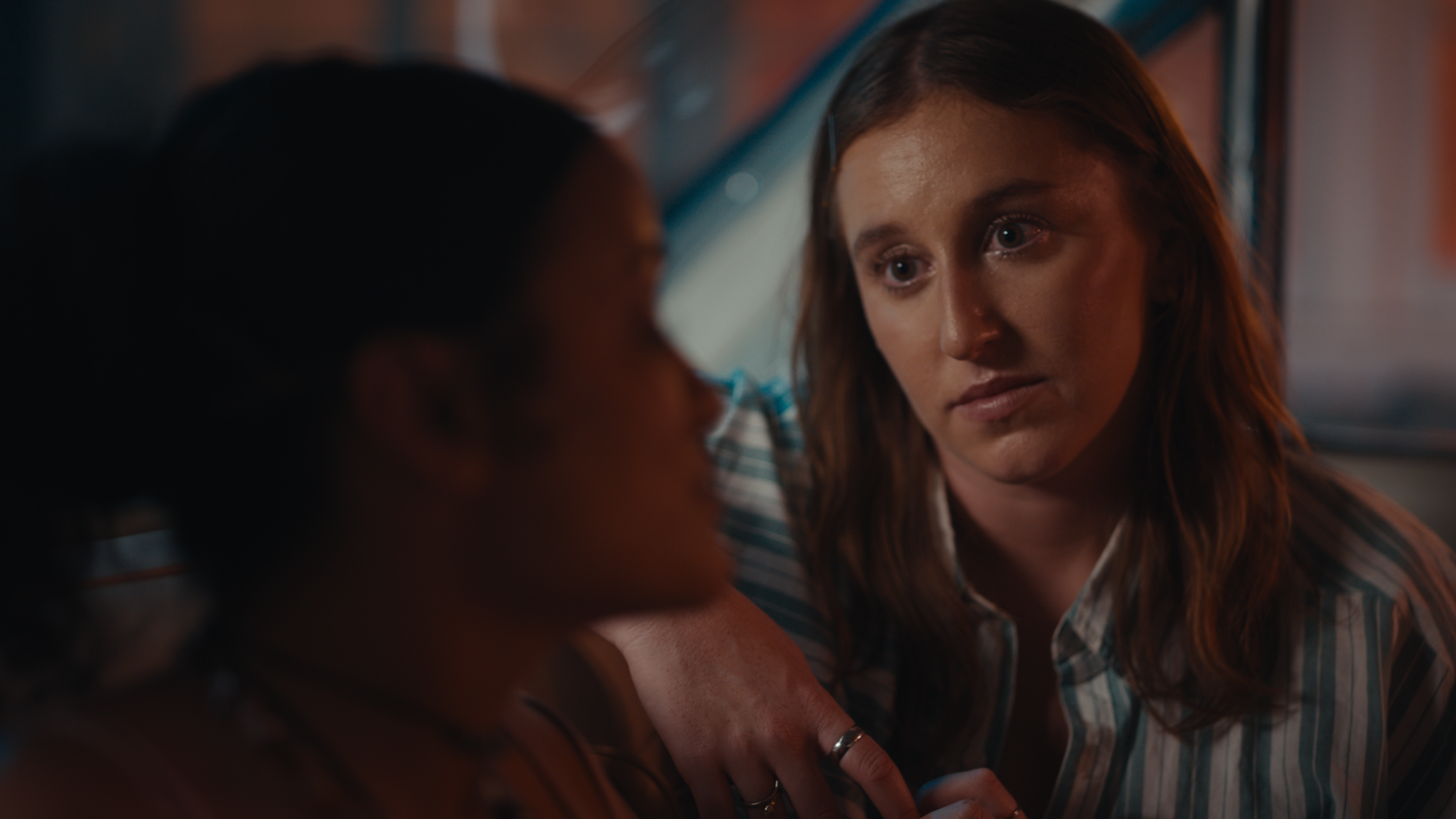 A woman with long brown hair and a striped shirt is looking intently at a person with dark curly hair, who is blurred in the foreground.