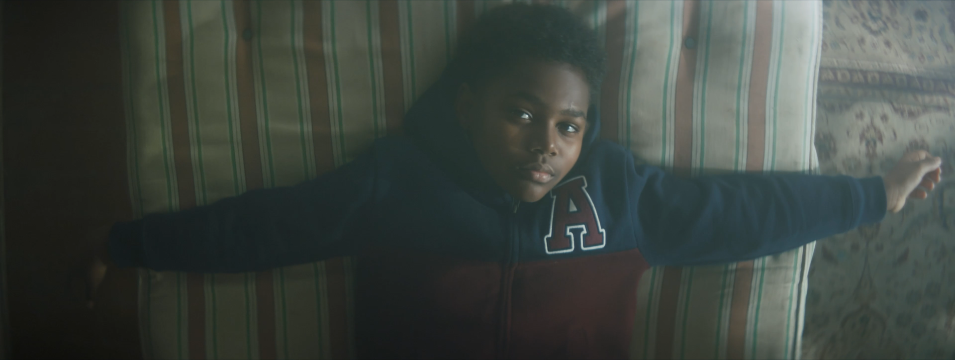 A young boy lying on a striped couch, looking up at the camera with a serious expression, wearing a navy blue hoodie with red and white letter patches.