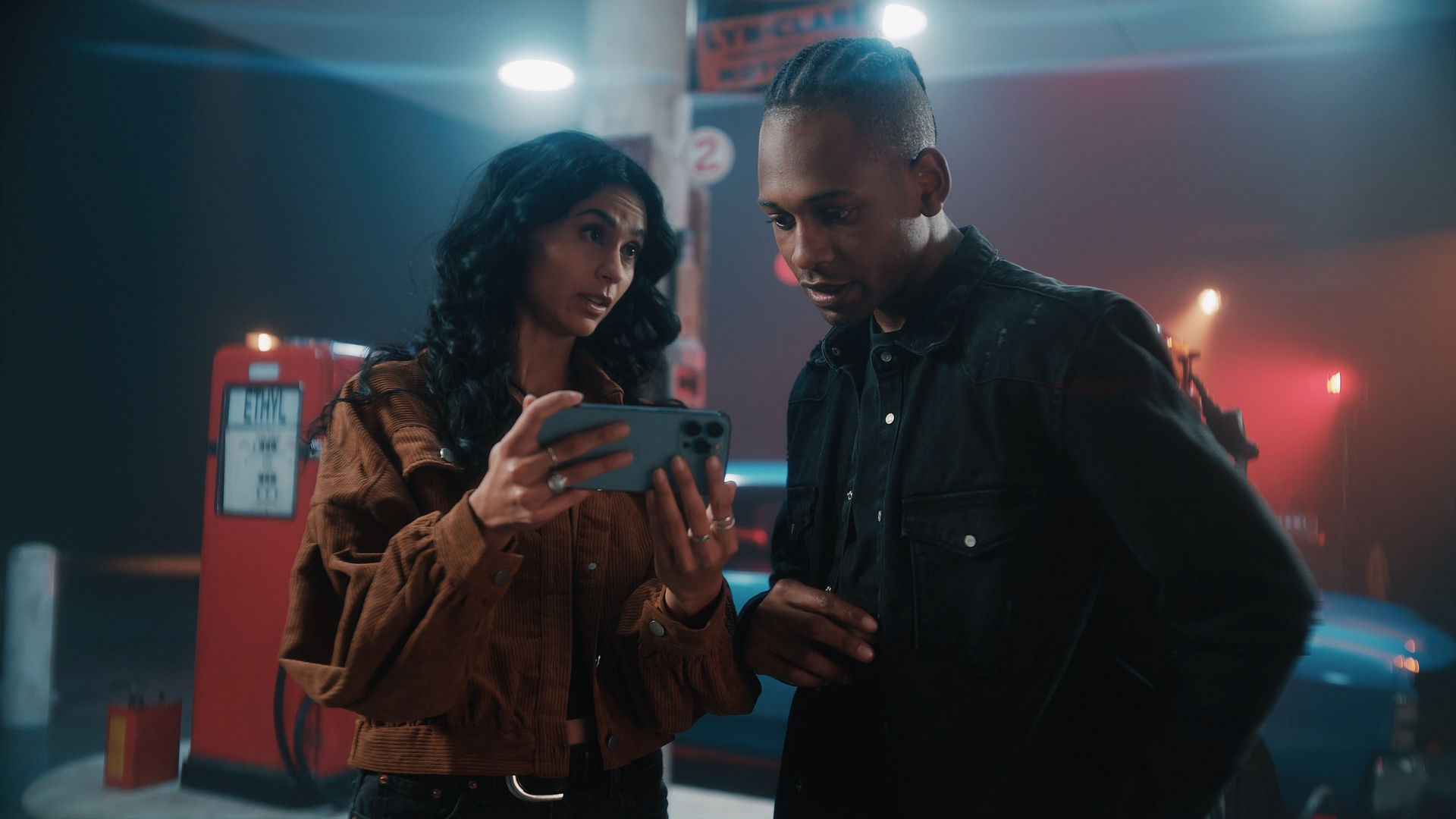 Two women, one holding a smartphone, are looking at the screen and conversing inside a dimly lit parking garage with red fire safety equipment in the background.