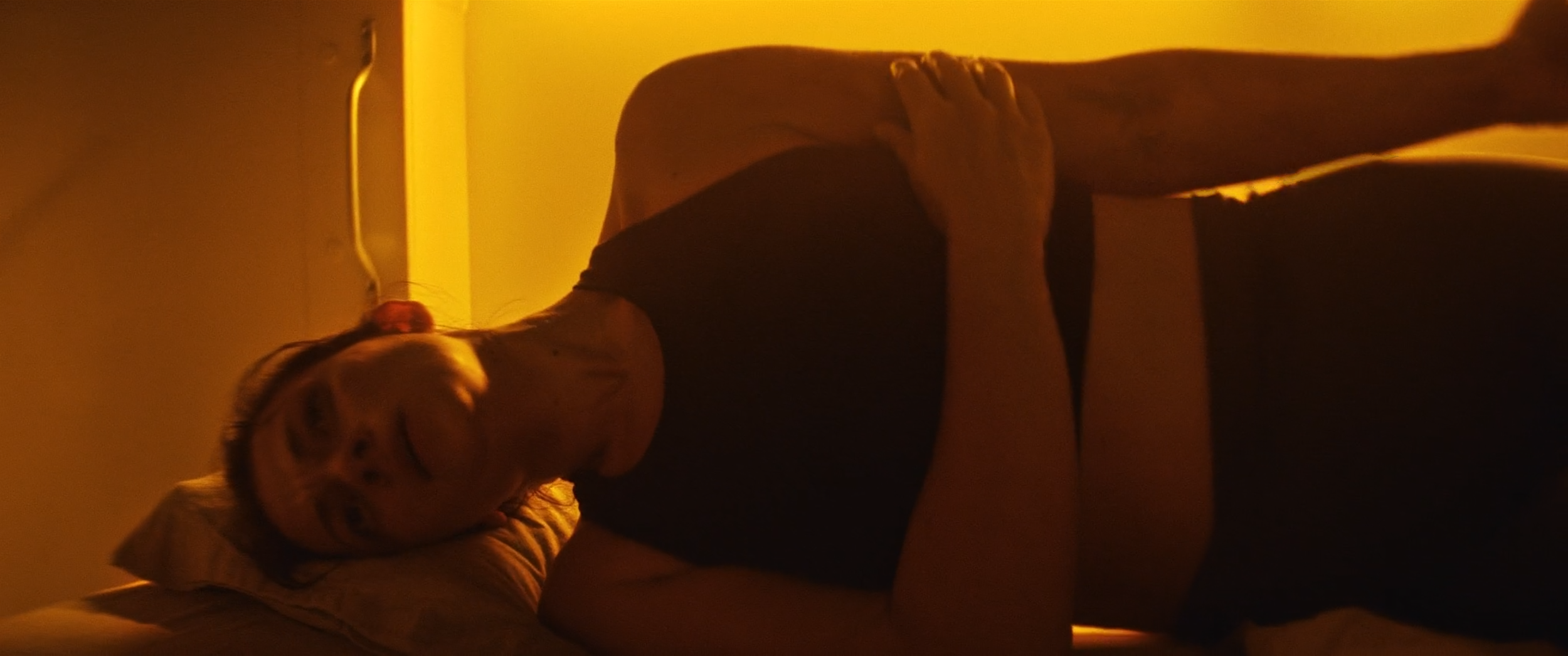 A woman lying on a bed, smiling with her eyes closed, in a dimly lit room with warm orange lighting.