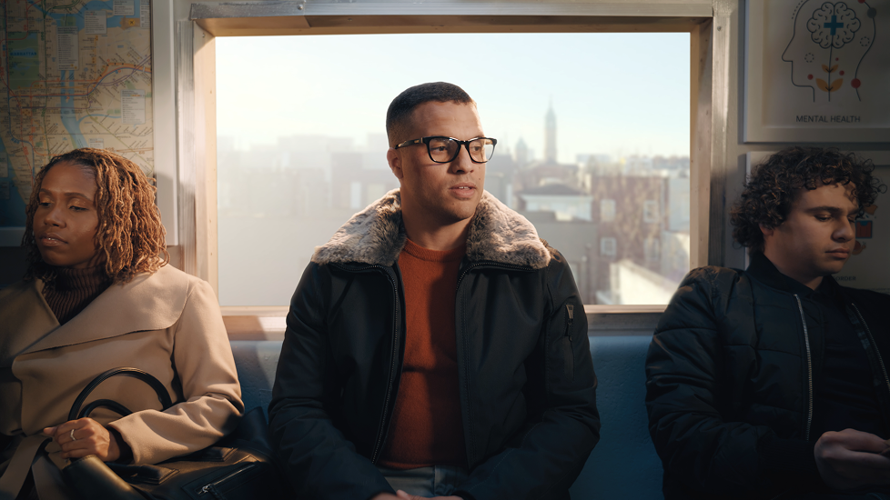 Three people sitting on a bus, looking away from the camera. A woman with curly hair and a tan coat is on the left, a man with glasses and a black jacket is in the middle, and another young man with curly hair and a dark jacket is on the right. City buildings are visible through the window behind them.