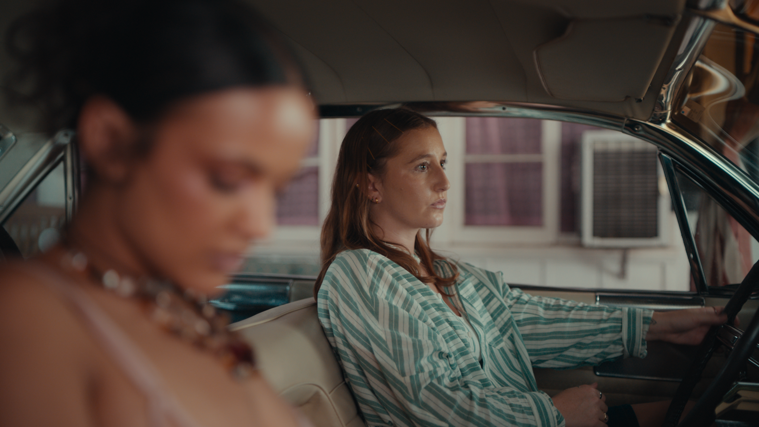 A woman with long reddish-brown hair in a striped shirt is driving a vintage car. A woman with dark hair and wearing jewelry is blurred in the foreground.