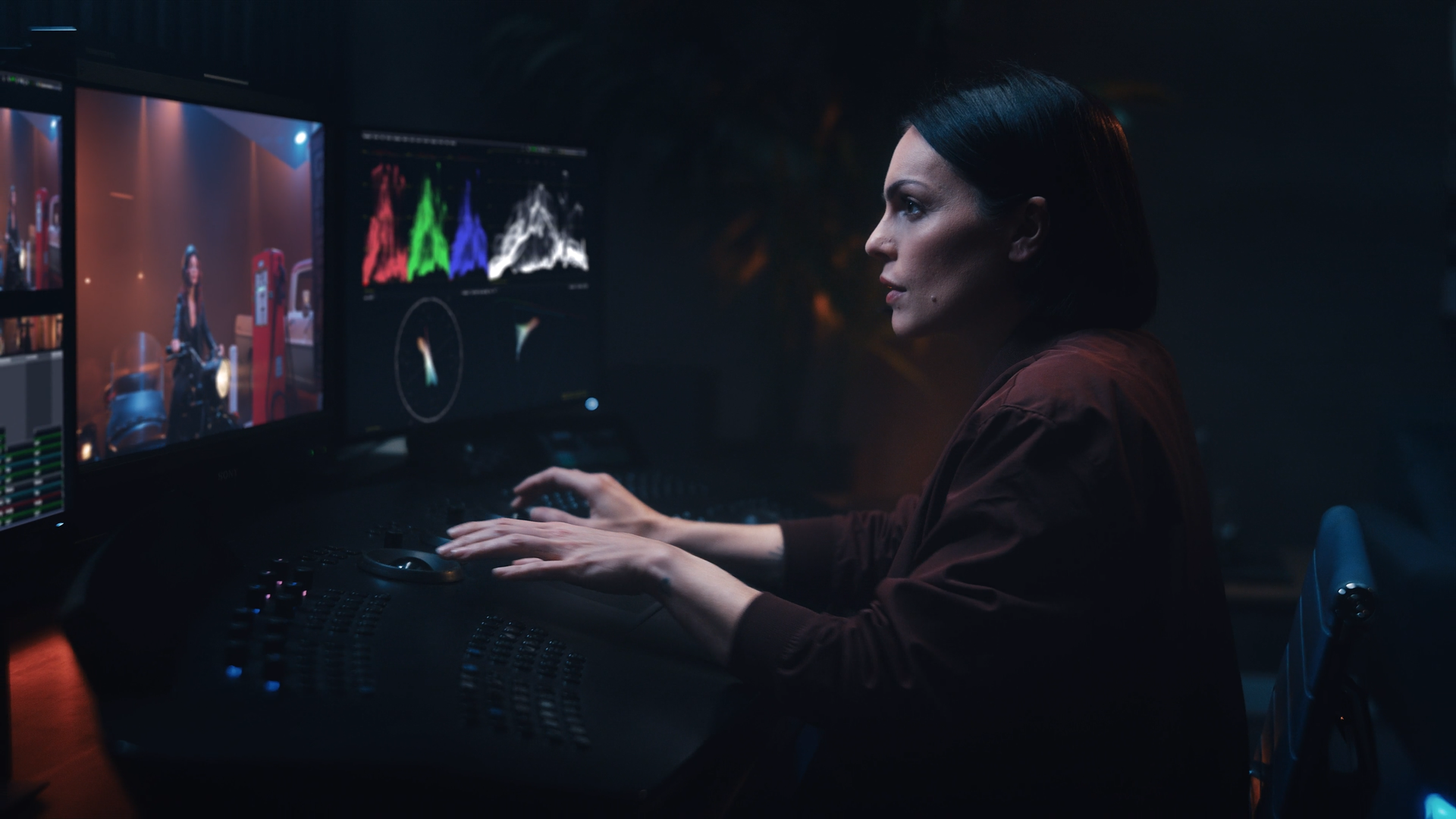 A woman working as a colorist in a dark editing studio, viewing multiple monitors displaying color correction and video editing software.