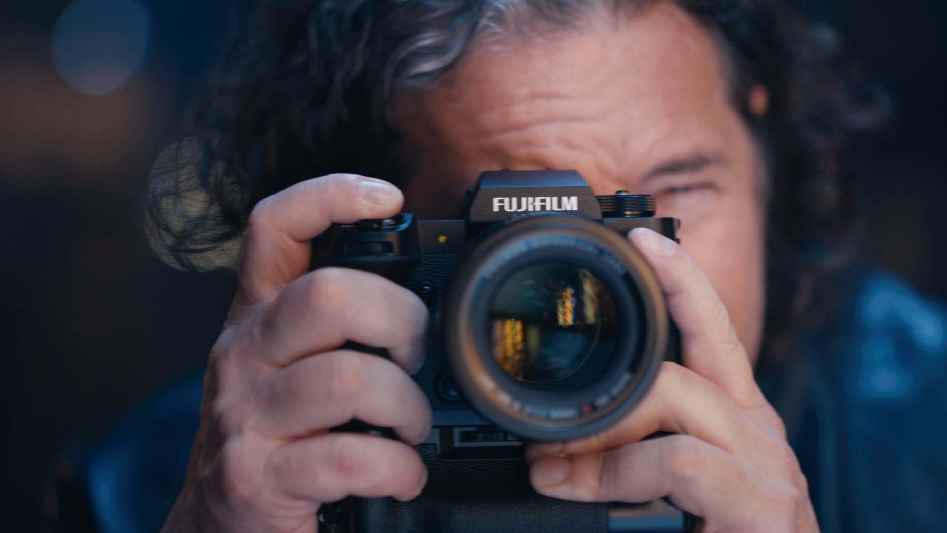 Person with curly hair holding a Fujifilm camera, taking a photo outdoors.