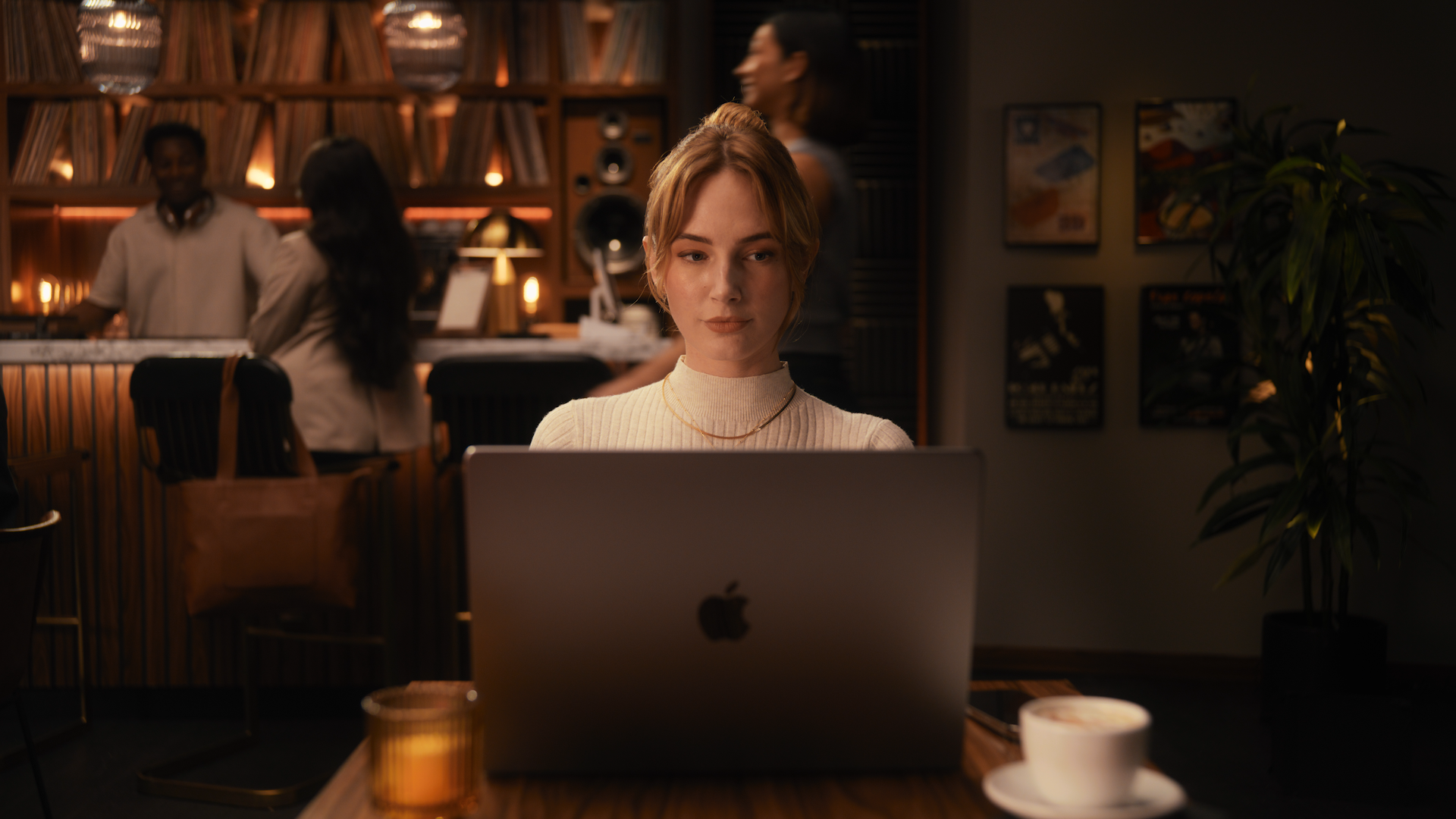 Woman with red hair working on a laptop in a cozy, dimly lit coffee shop or cafe with a bookshelf and people in the background.