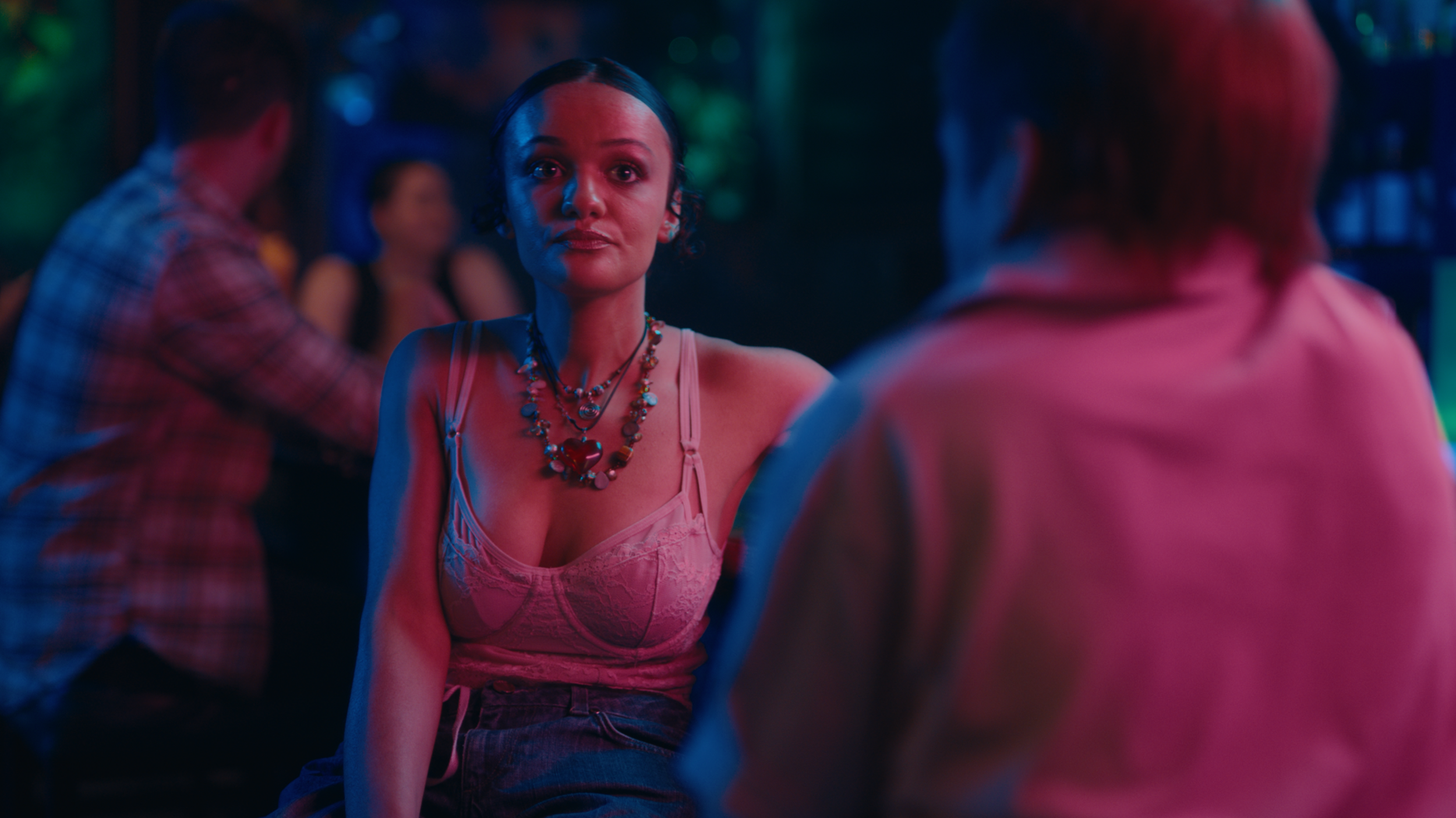 A woman with dark hair and a pink lace top, wearing colorful necklaces and earrings, is at a party or club with multicolored lighting. She is looking forward with a neutral or slightly serious expression, surrounded by other people who are out of focus.