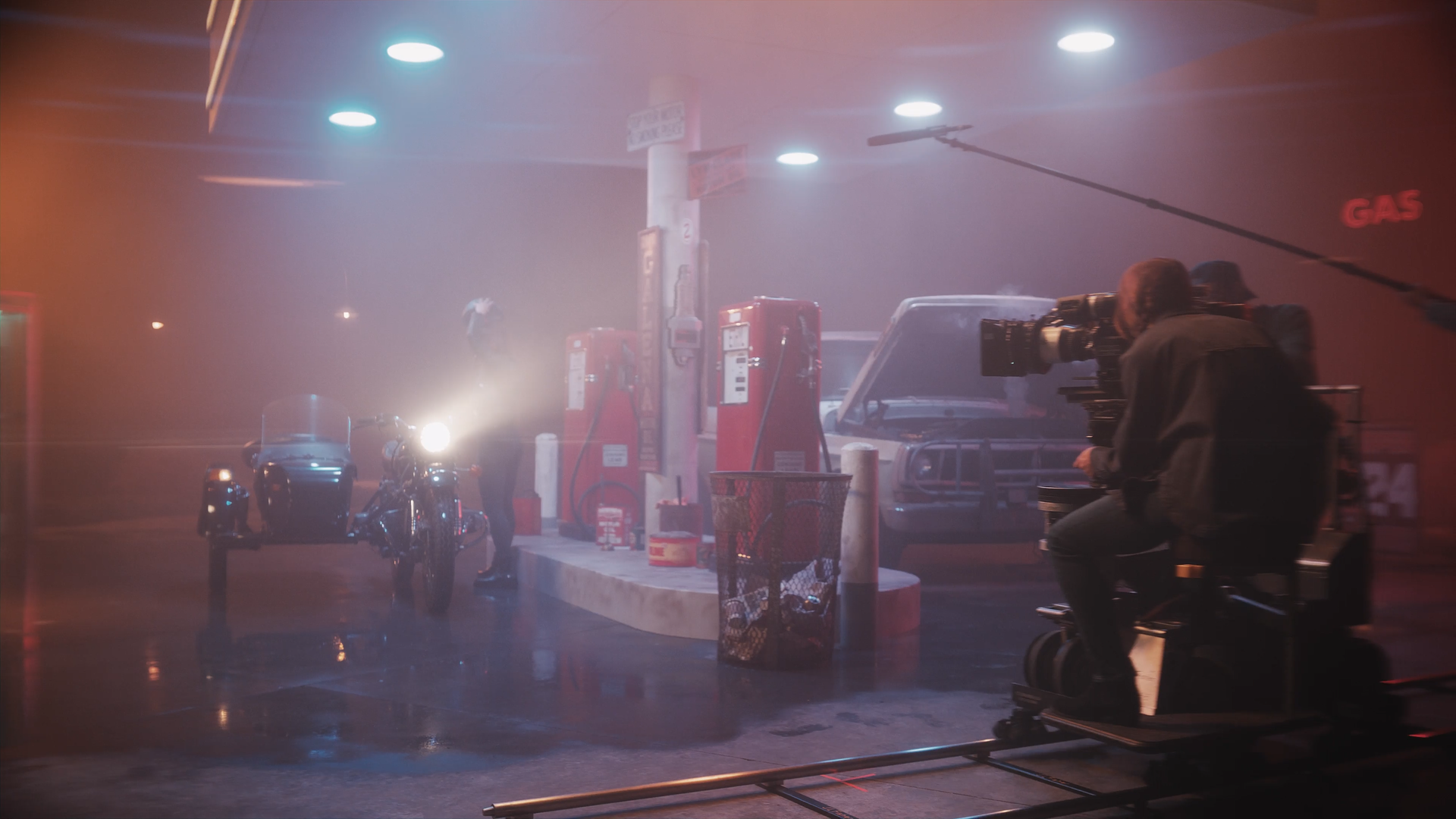 Nighttime scene at a gas station with motorcycles, a car, and filming equipment including a camera operator with a camera mounted on a stabilizer. The gas station has red fuel pumps and overhead lights, with fog or smoke present.