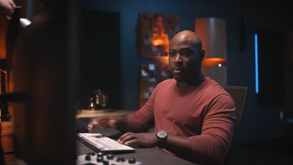 A man in a reddish-brown shirt sitting at a studio or control room desk looking at a monitor with a focused expression.
