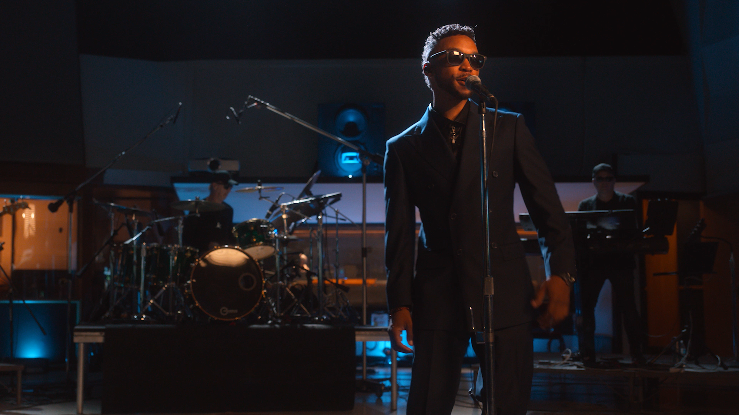 Male singer wearing sunglasses and a black suit performing on stage with a microphone, accompanied by a band including a drummer and keyboardist, in a dimly lit recording studio or concert hall.