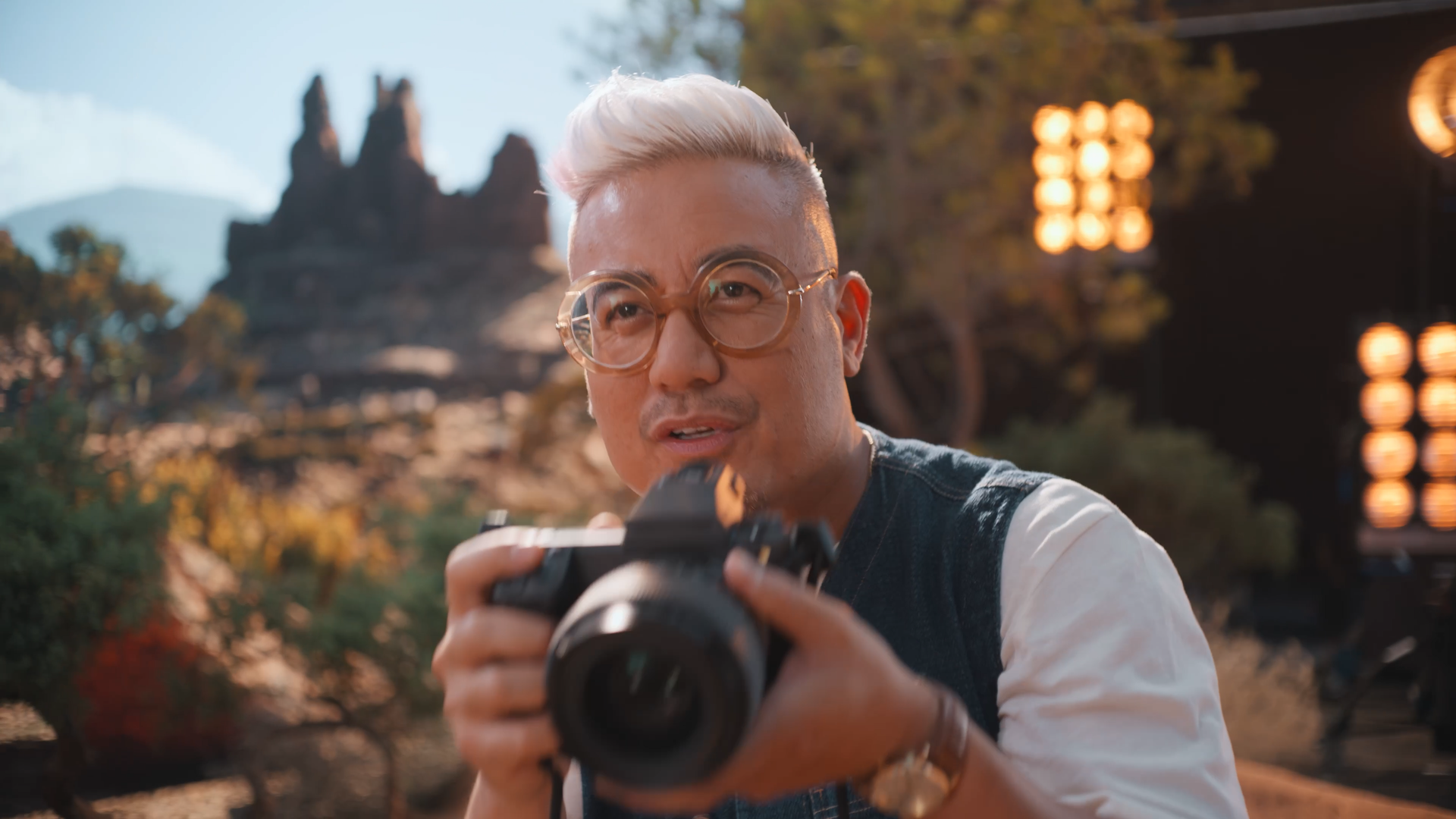 Man with platinum blonde hair and glasses holding a camera outdoors in a park or garden setting, with trees and a mountain in the background and warm lighting effects.
