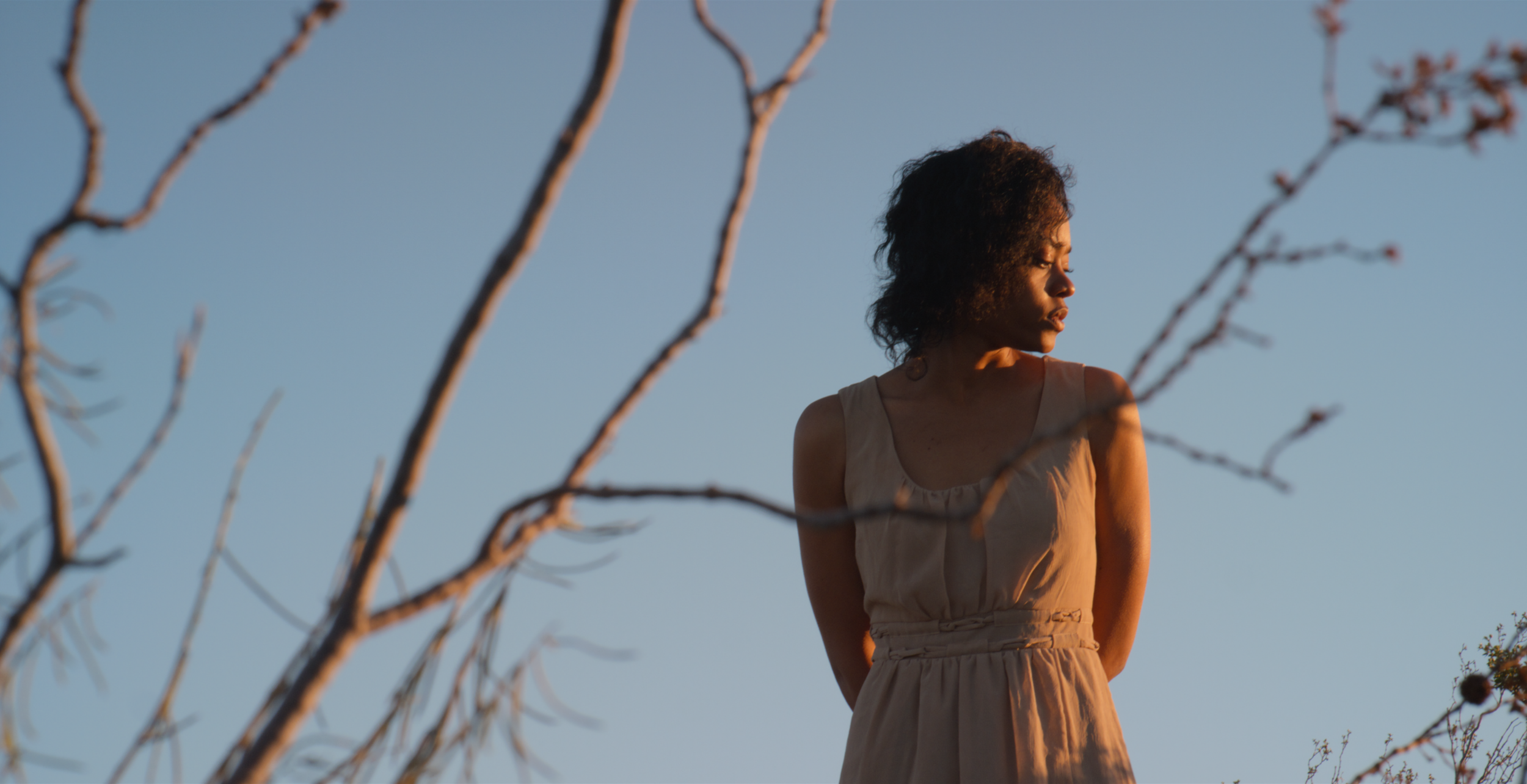 A woman with curly hair and dark skin standing outdoors against a clear blue sky, framed by leafless branches in the foreground, wearing a sleeveless beige dress, looking to her right.