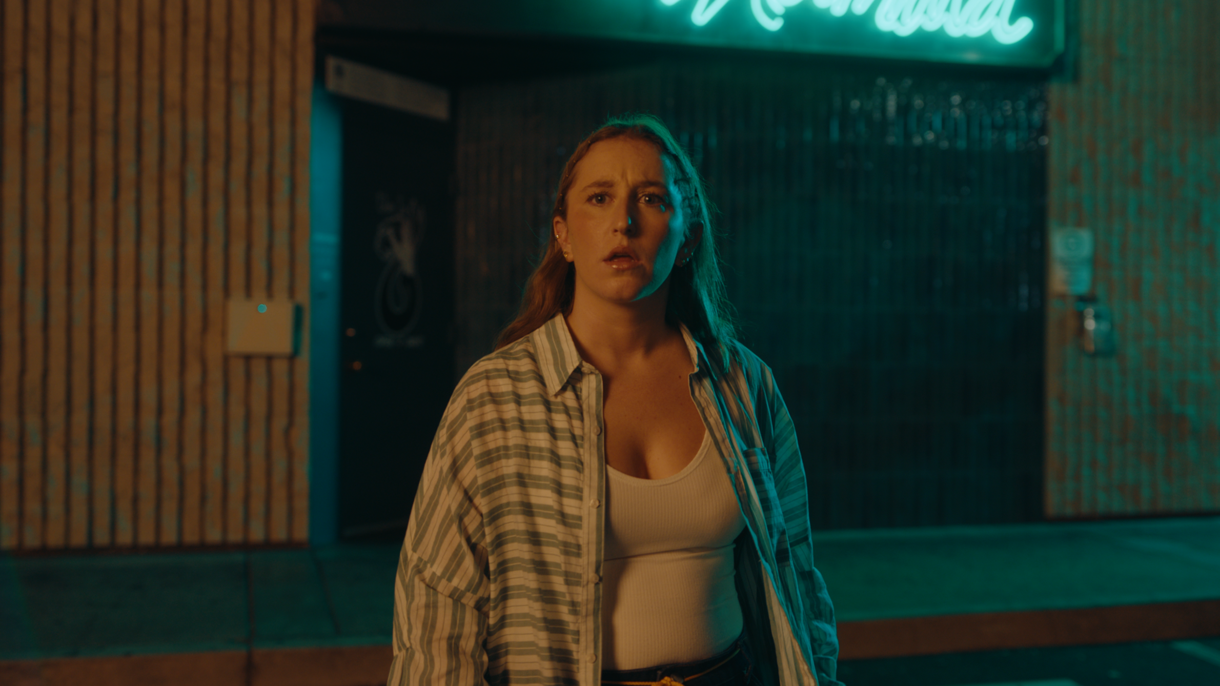 A woman with long hair looking surprised or shocked, standing outside a building at night with neon signs and a wooden wall in the background.