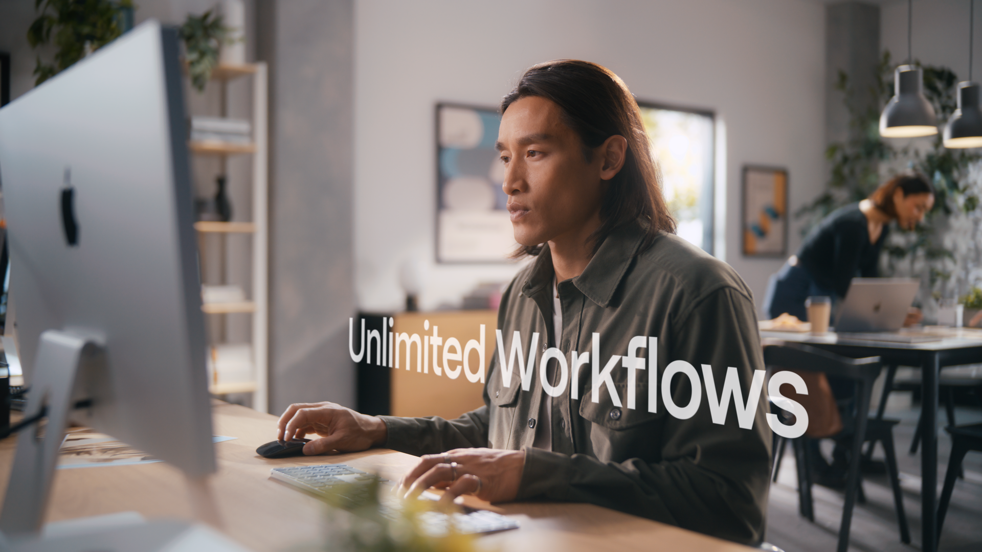 A man working at a computer in an office with two women in the background, one using a laptop and the other standing, with natural light coming through a window and the text 'Unlimited Workflows' overlaid.
