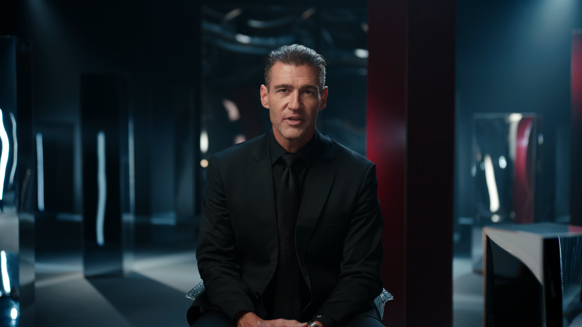 A man dressed in a black suit sitting in a dark, modern room with neon lights and futuristic decor.