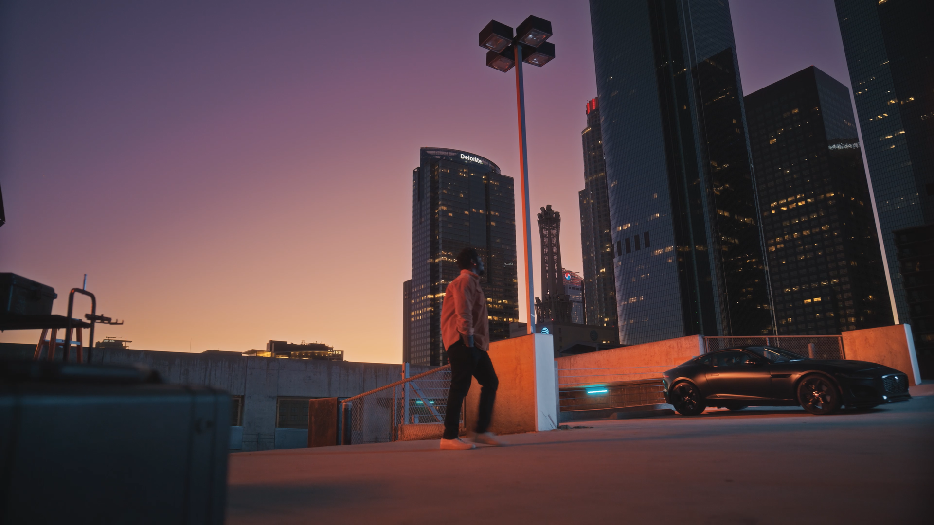 Man walking on rooftop parking garage with a luxury black sports car, tall city buildings with illuminated windows, and a sunset sky in the background.