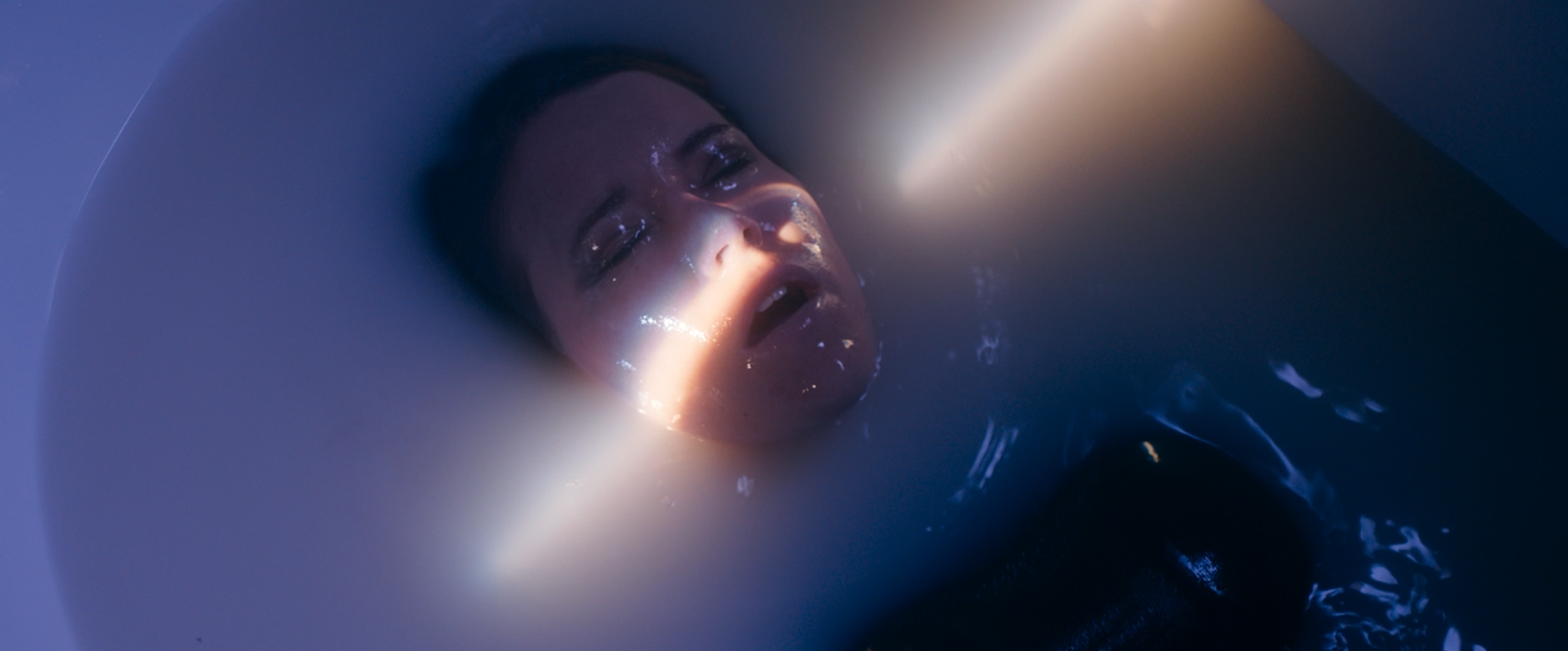 Person with long hair submerged in a bathtub with water, eyes closed, water reflecting light, and face partially visible.