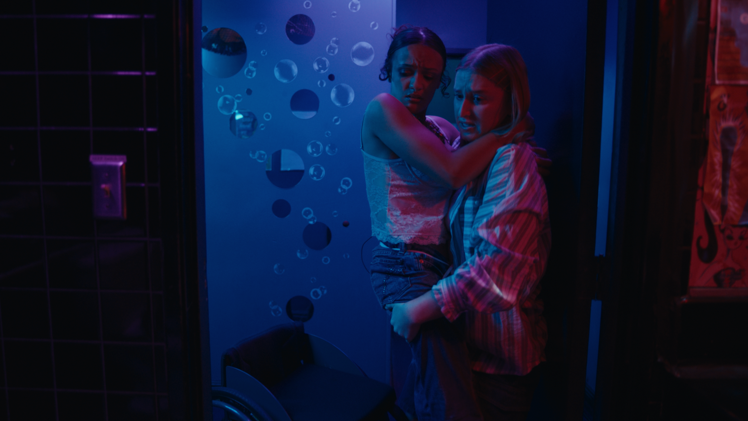 Two women are embracing in a dimly lit room with blue and purple lighting, against a wall decorated with circular mirrors or reflective shapes.