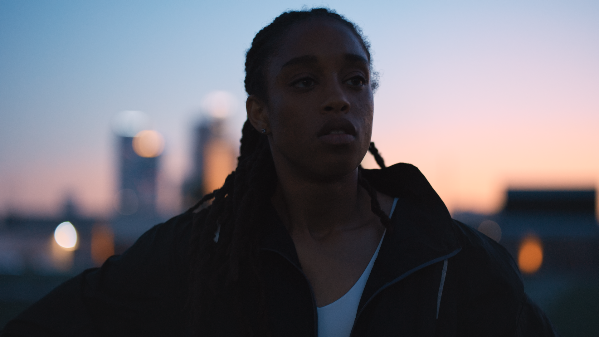 A woman with dark braided hair wearing a black jacket and white top, standing outdoors during sunset with a city skyline in the background.