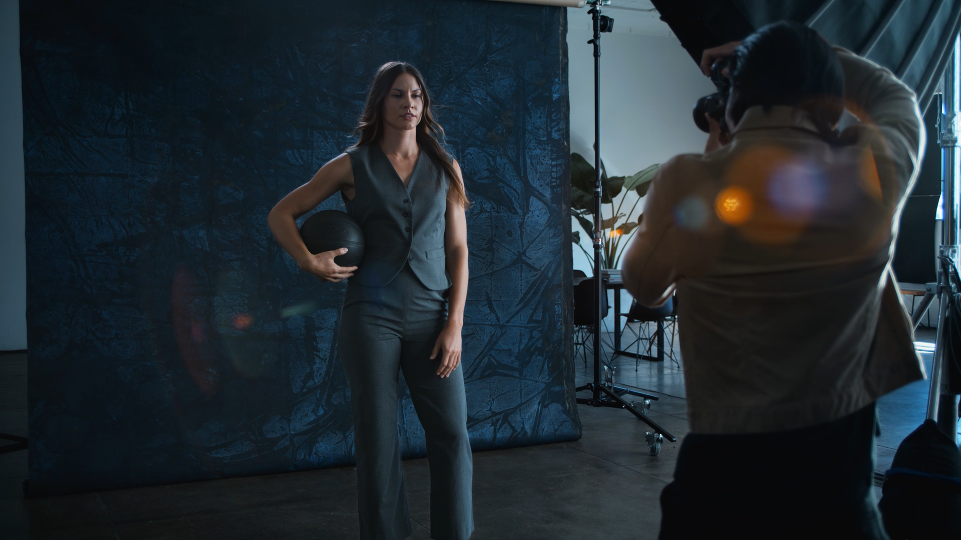 A professional woman in a gray suit holding a black medicine ball during a photo shoot with a photographer taking pictures in a studio setting.