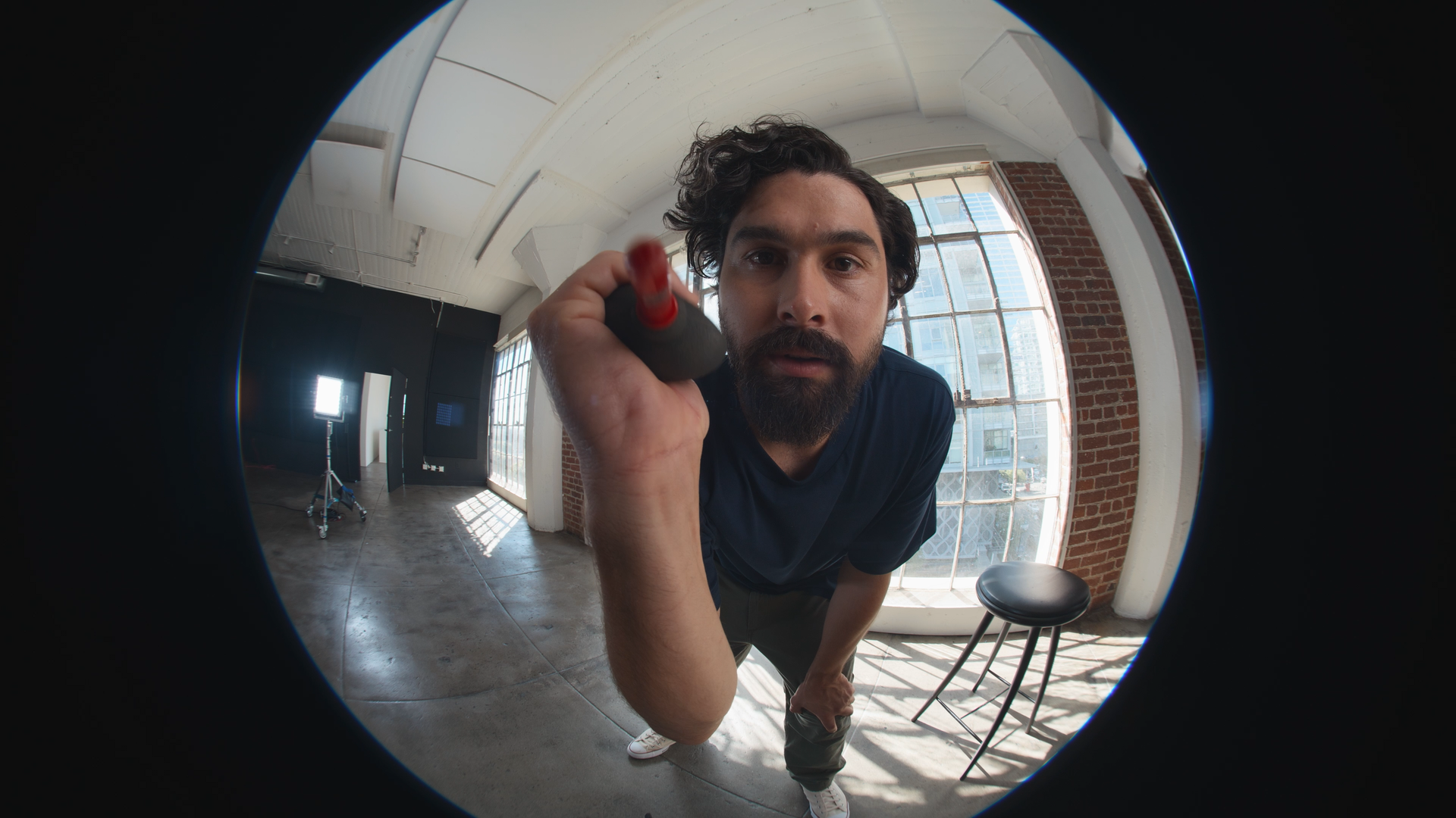Man with curly hair and beard taking a selfie with a wide-angle lens in a studio with large windows, brick wall, and studio equipment.