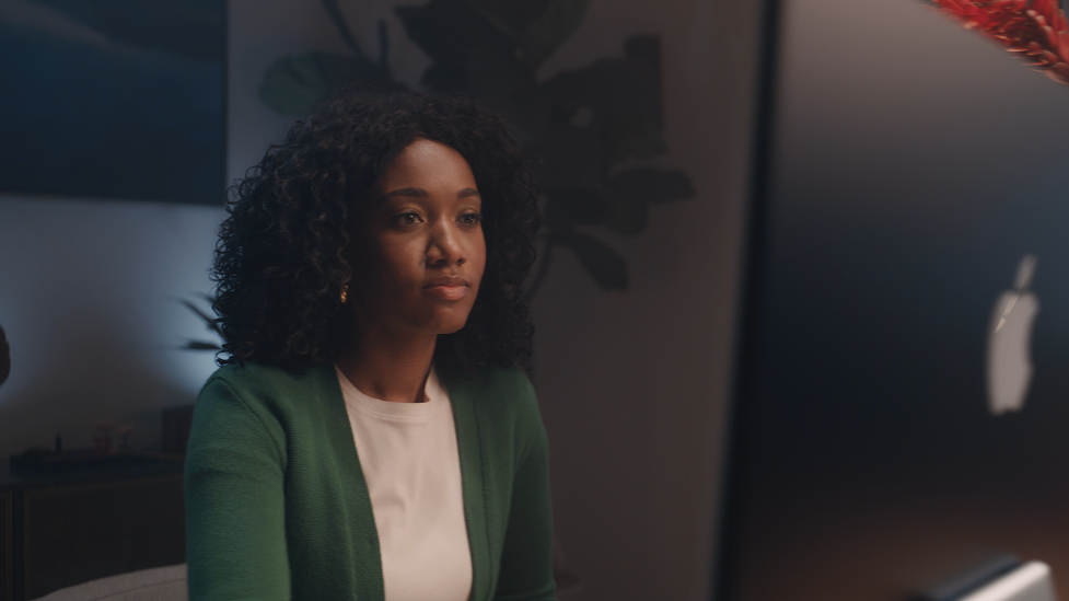 A woman with curly hair and a green cardigan sitting at a desk in front of a computer monitor.