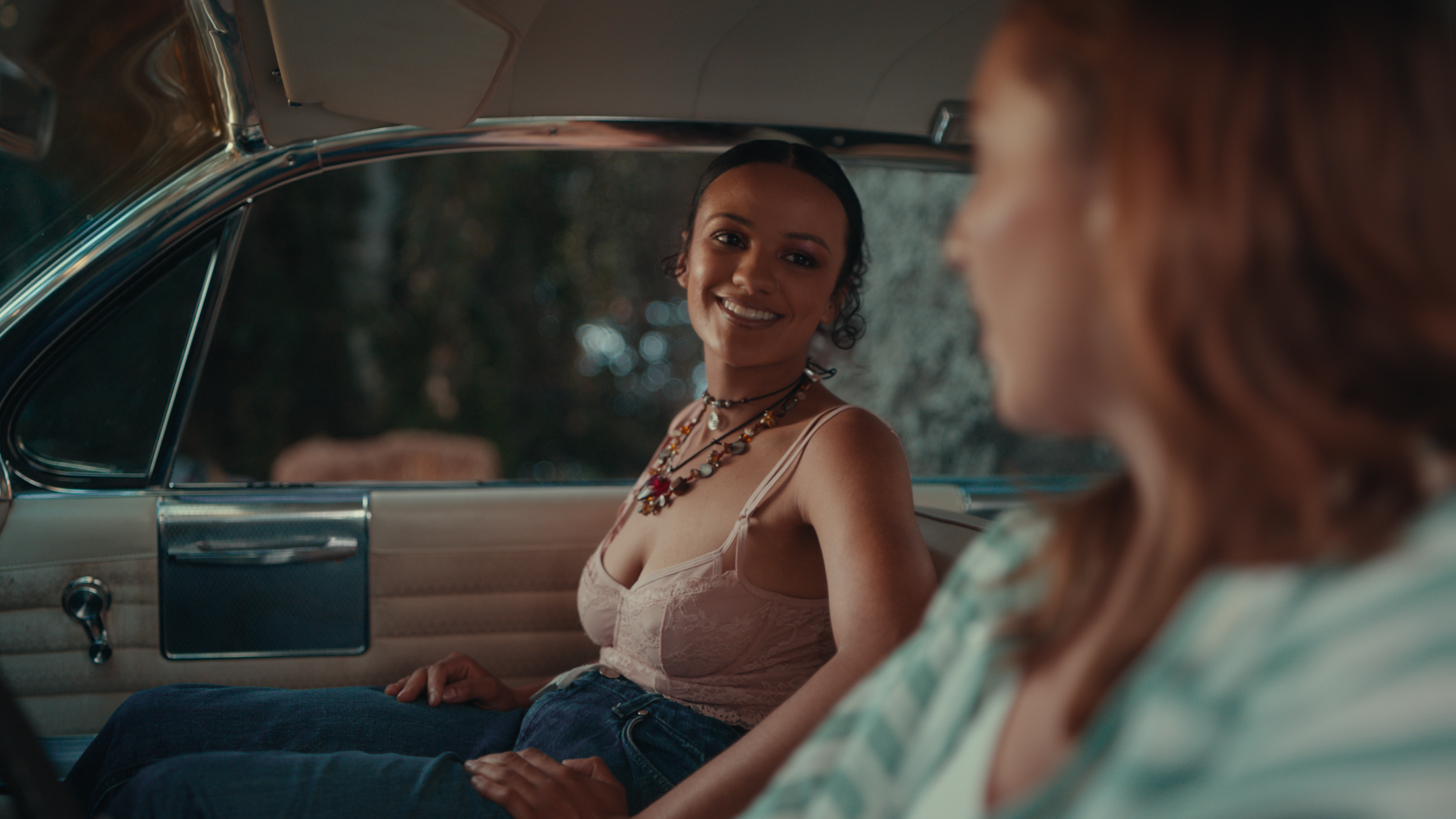Two women sitting in a vintage car, one woman with dark hair and a necklace smiling at the other woman with red hair, seen from behind.