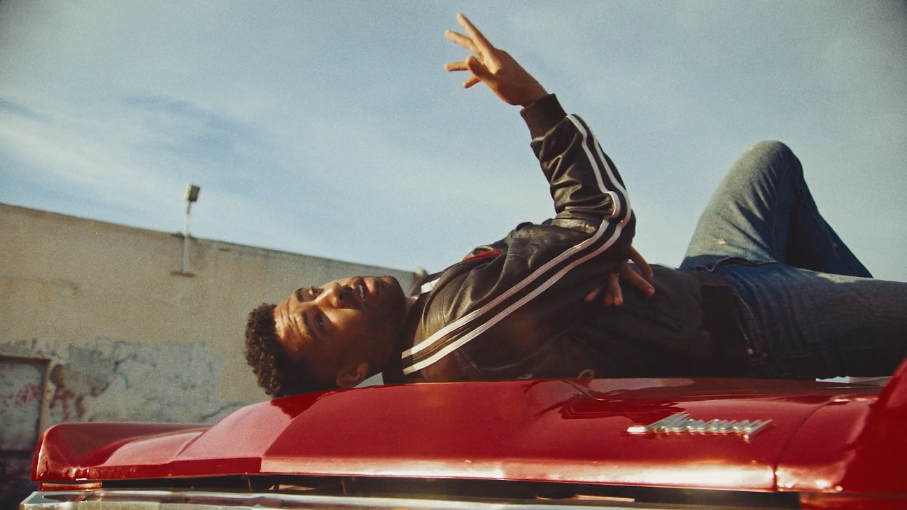 A man lying on the hood of a red car, looking at the sky with one hand raised and making a peace sign. The car emblem reads 'Corvette'.