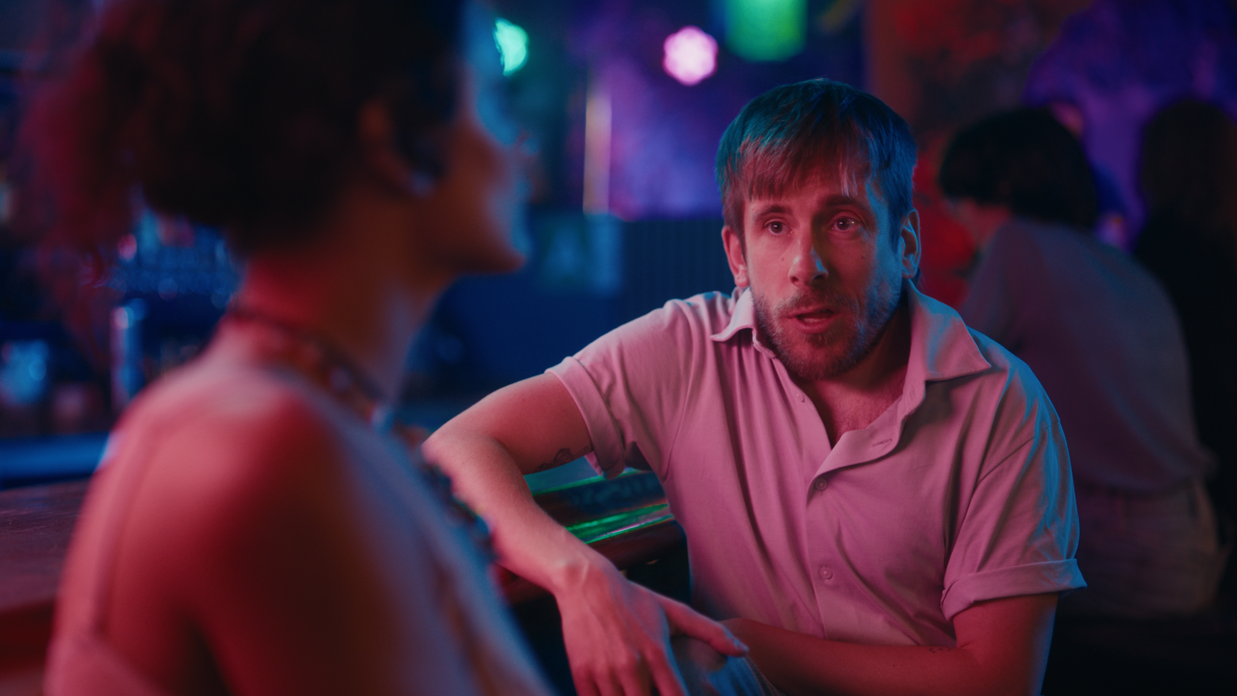 A man with light brown hair and a beard wearing a light pink shirt is talking to a woman with curly hair in a dimly lit bar or nightclub with colorful neon lights.
