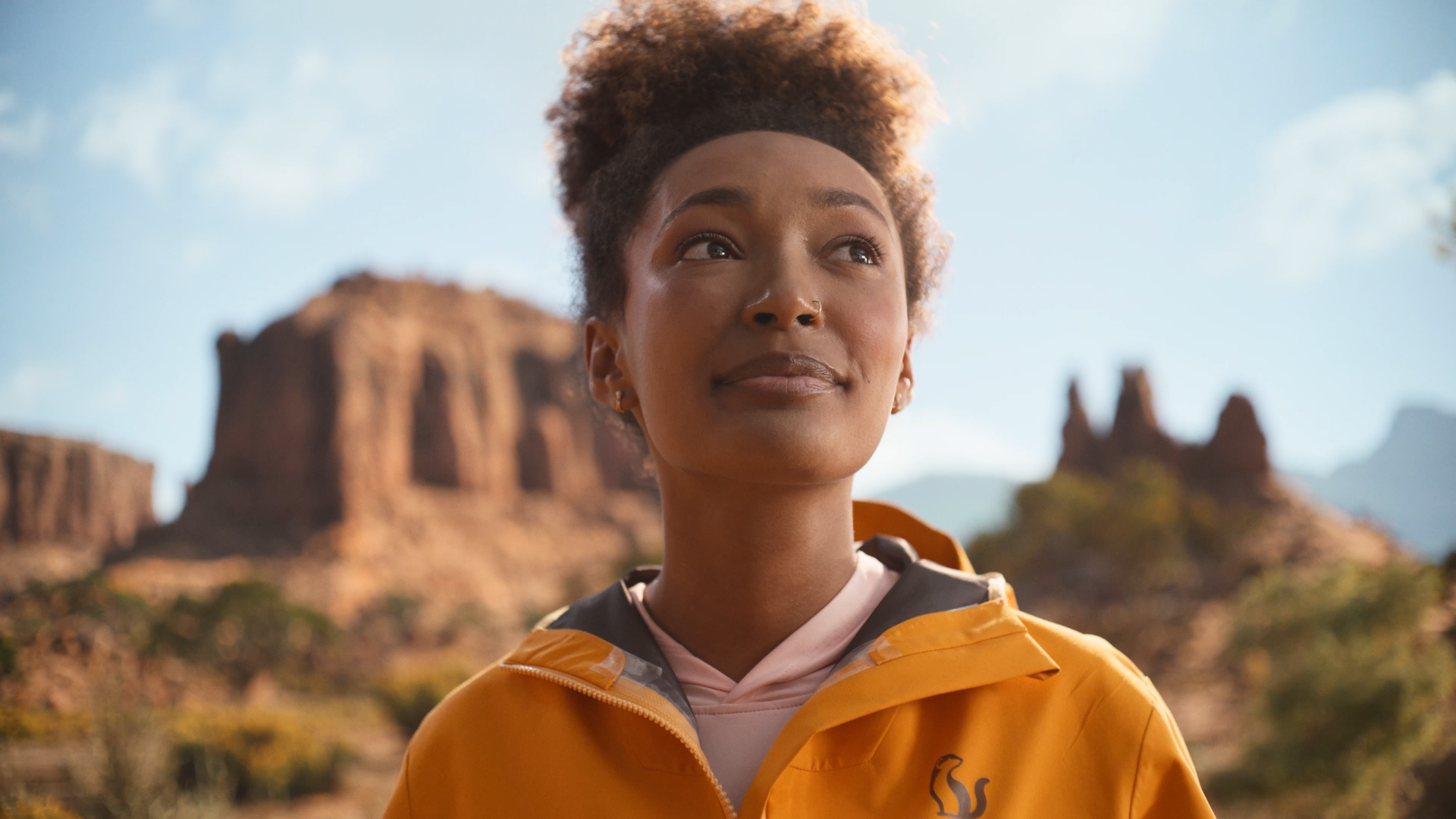 A woman with short curly hair wearing a yellow rain jacket outdoors against a desert landscape with large rock formations, blue sky, and clouds.