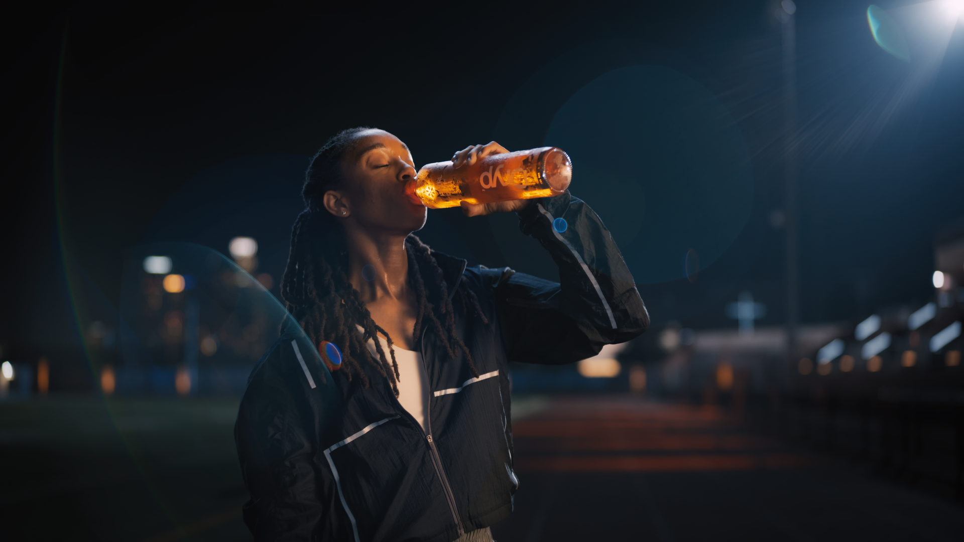 A woman with dreadlocks wearing a black jacket drinking from a sports drink bottle at night in an outdoor sports setting.