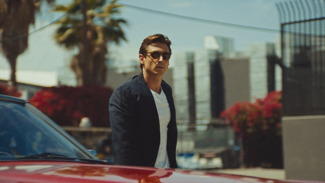 A man wearing sunglasses, a black jacket, and a striped shirt stands next to a red car outdoors with palm trees and buildings in the background.