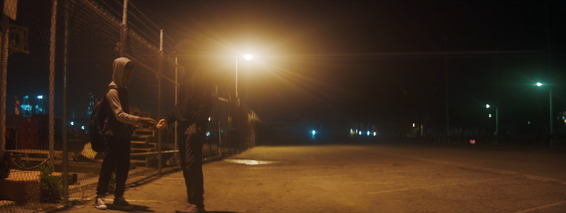 Two young people exchanging something at night near a chain-link fence, illuminated by streetlights.