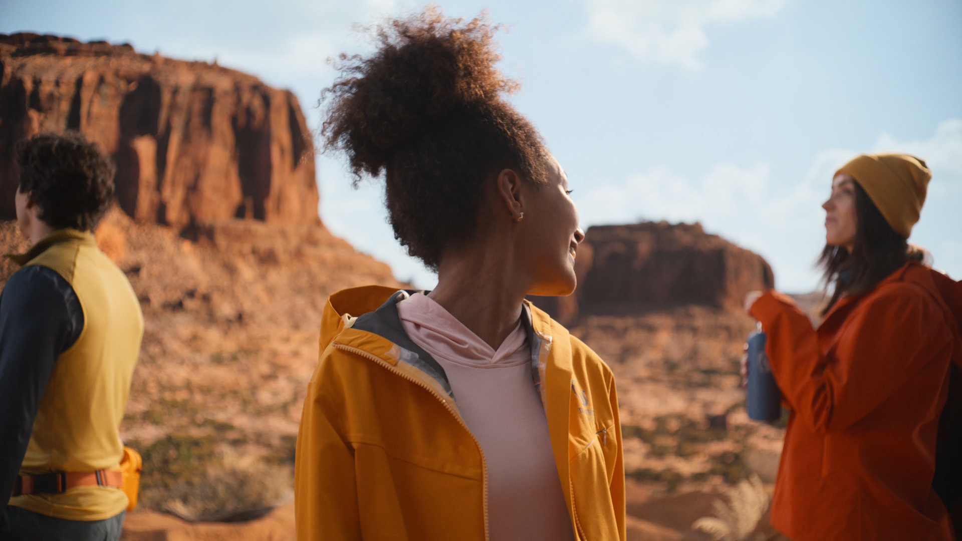 Three women standing outdoors in a rocky desert landscape, smiling and talking to each other during daytime.